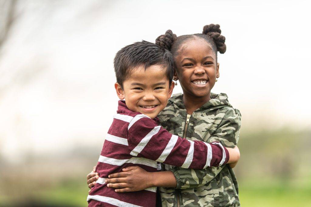 A young Asian boy and a sweet African girl warmly hug each other while outside on a cool fall day. They are each dressed casually in autumn clothing and smiling as they enjoy their time together and the fresh air.